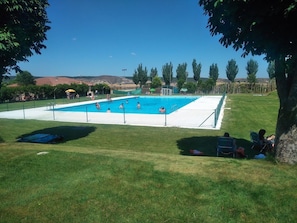 Una piscina rectangular envoltada per un terrat blanc, amb una zona enjardinada al primer terme i arbres al fons.