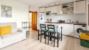 Een compact woongedeelte met een kitchenette, een eethoek met tafel en stoelen, een bank met een geel kussen, en een wasmachine.