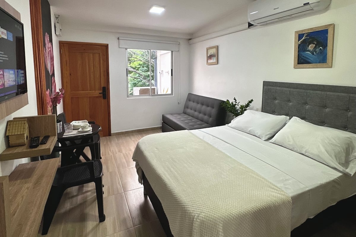 A bedroom with a bed, a desk, a chair, a window, and an air conditioning unit.