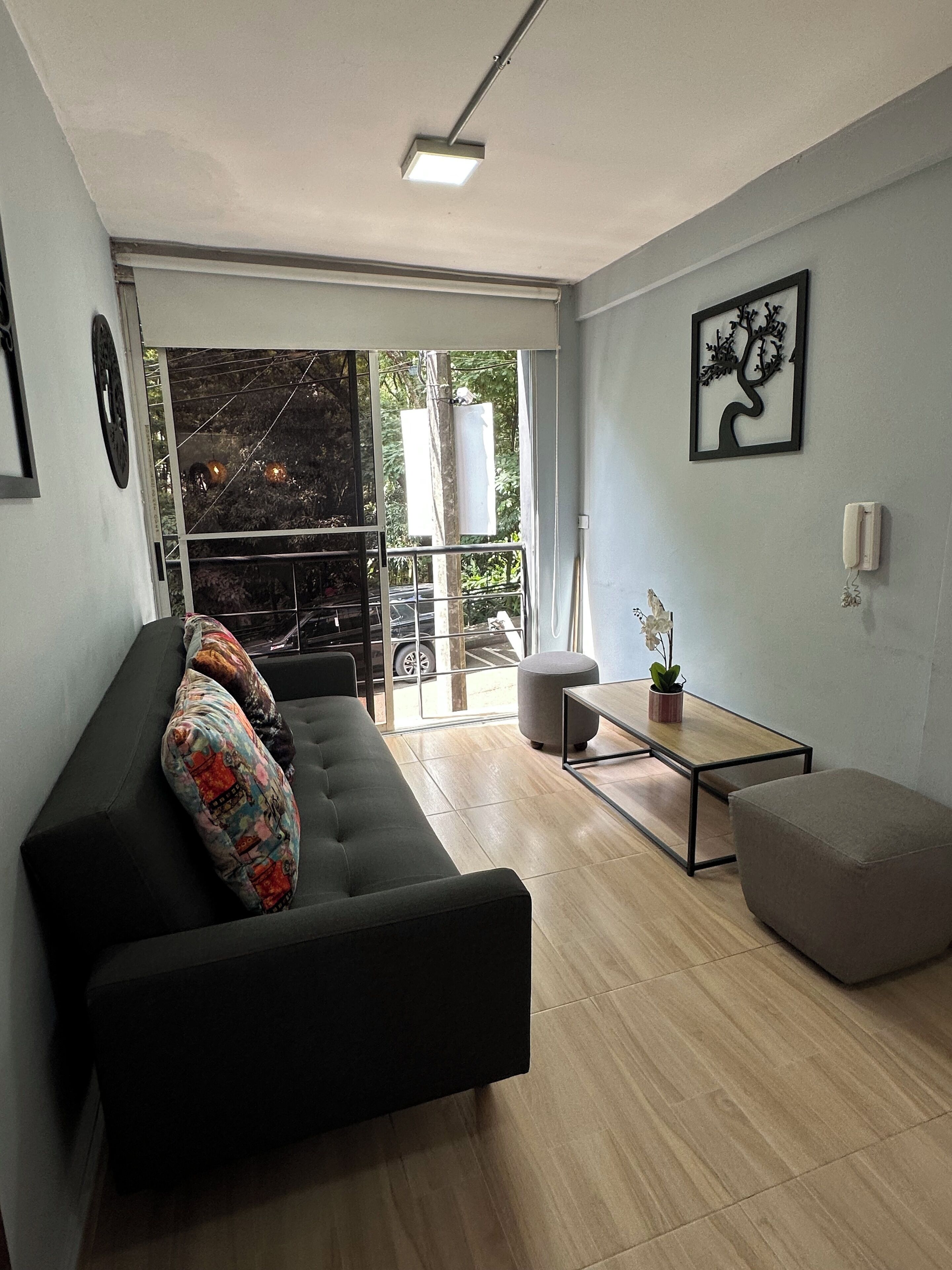 A modern living room with a black sofa, a small table, and a grey ottoman. There is a large window with a view of the outside.