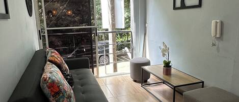 A modern living room with a black sofa, a small table, and a grey ottoman. There is a large window with a view of the outside.