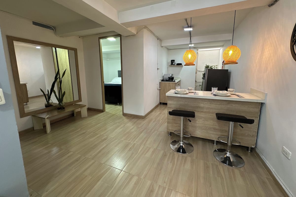 A modern kitchen with a central island, bar stools, pendant lighting, and a visible doorway leading to a bedroom.