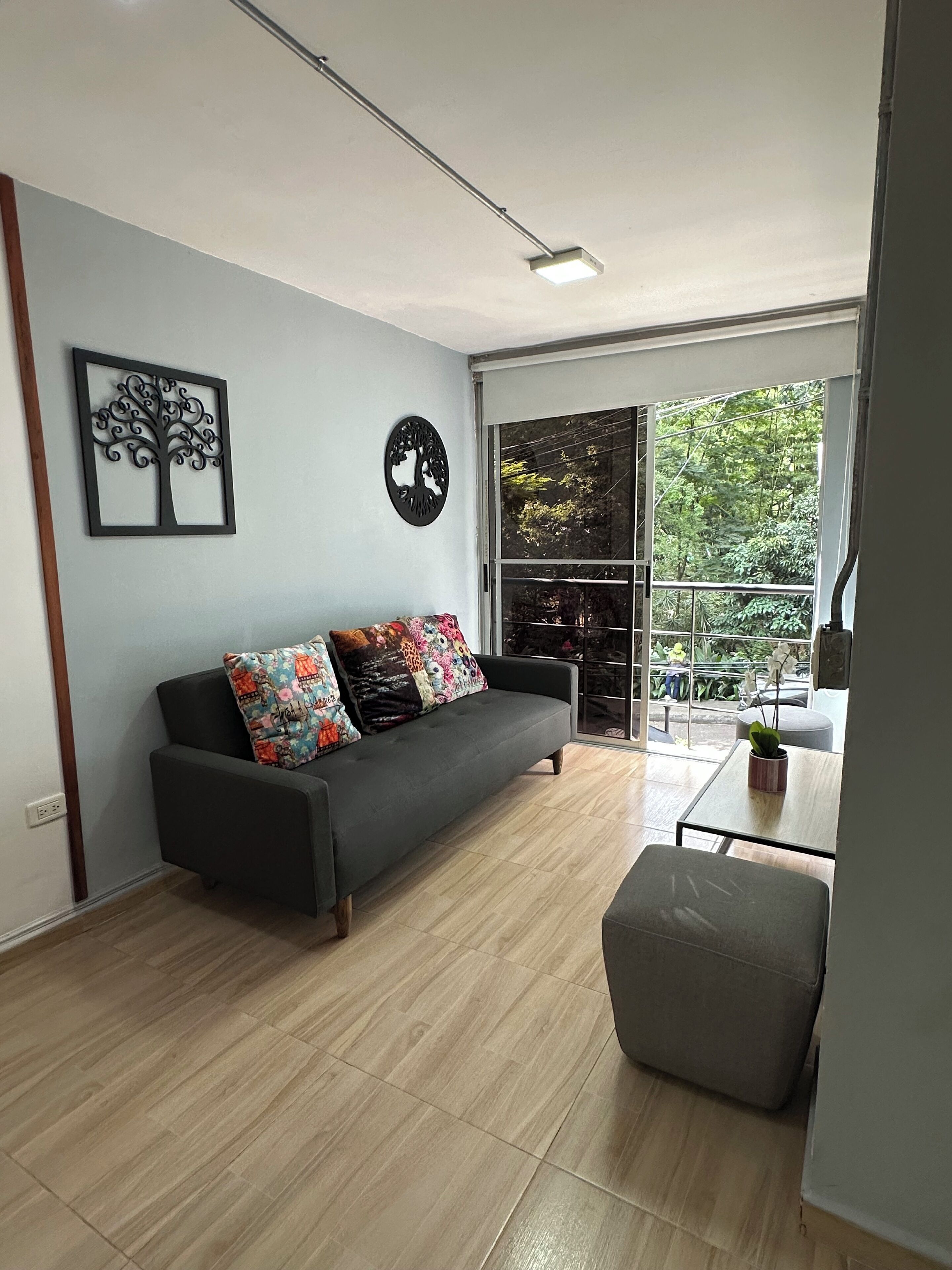 A modern living room with a grey sofa, colorful cushions, a small table with a plant, and a large window offering a view of greenery outside.