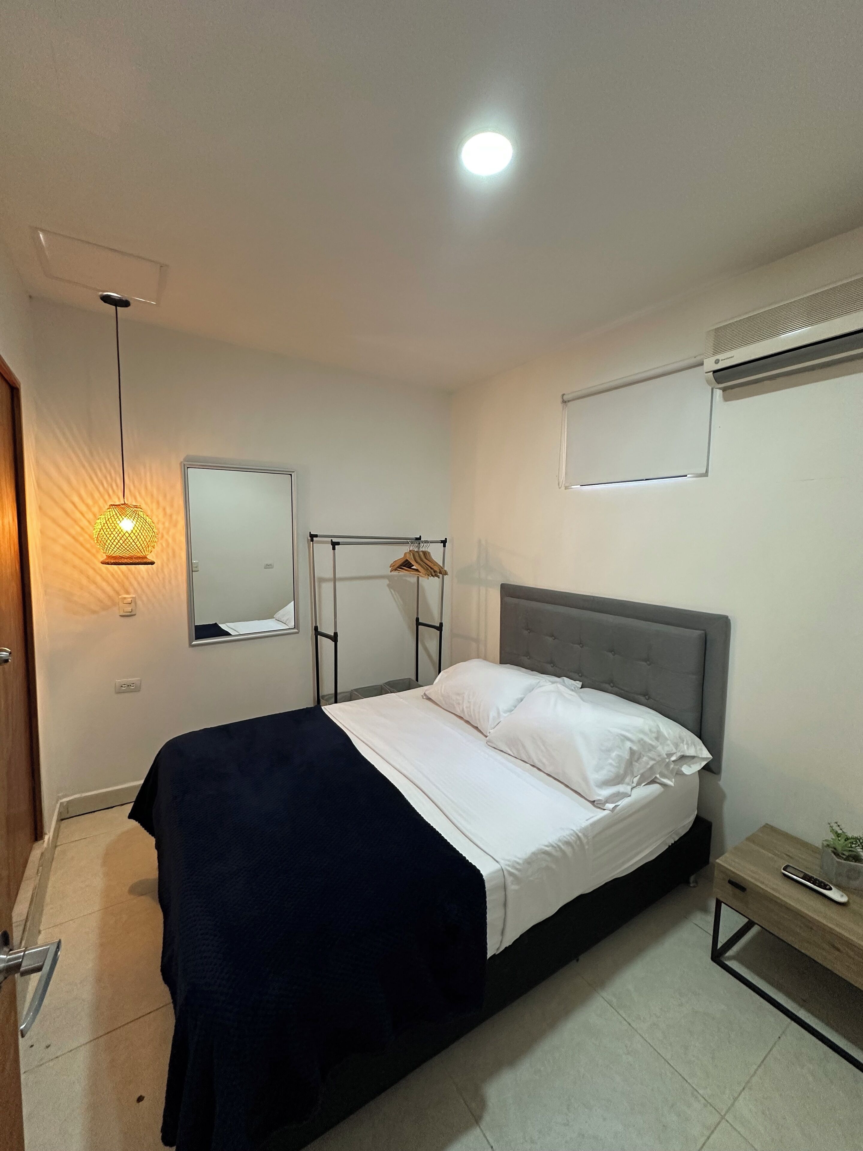 A bedroom with a bed, a bedside table, a hanging light fixture, a wall-mounted air conditioner, and a door.