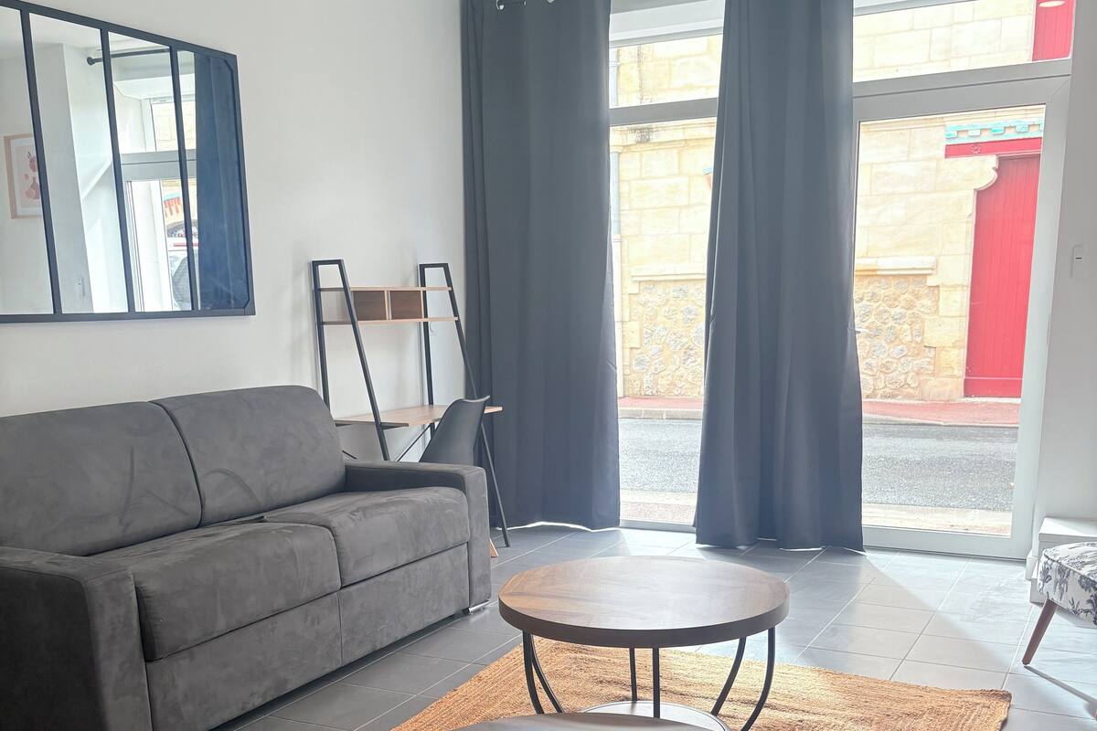 Un salon moderne avec un canapé gris, deux petites tables rondes, une table d’appoint en bois et un grand miroir au mur.