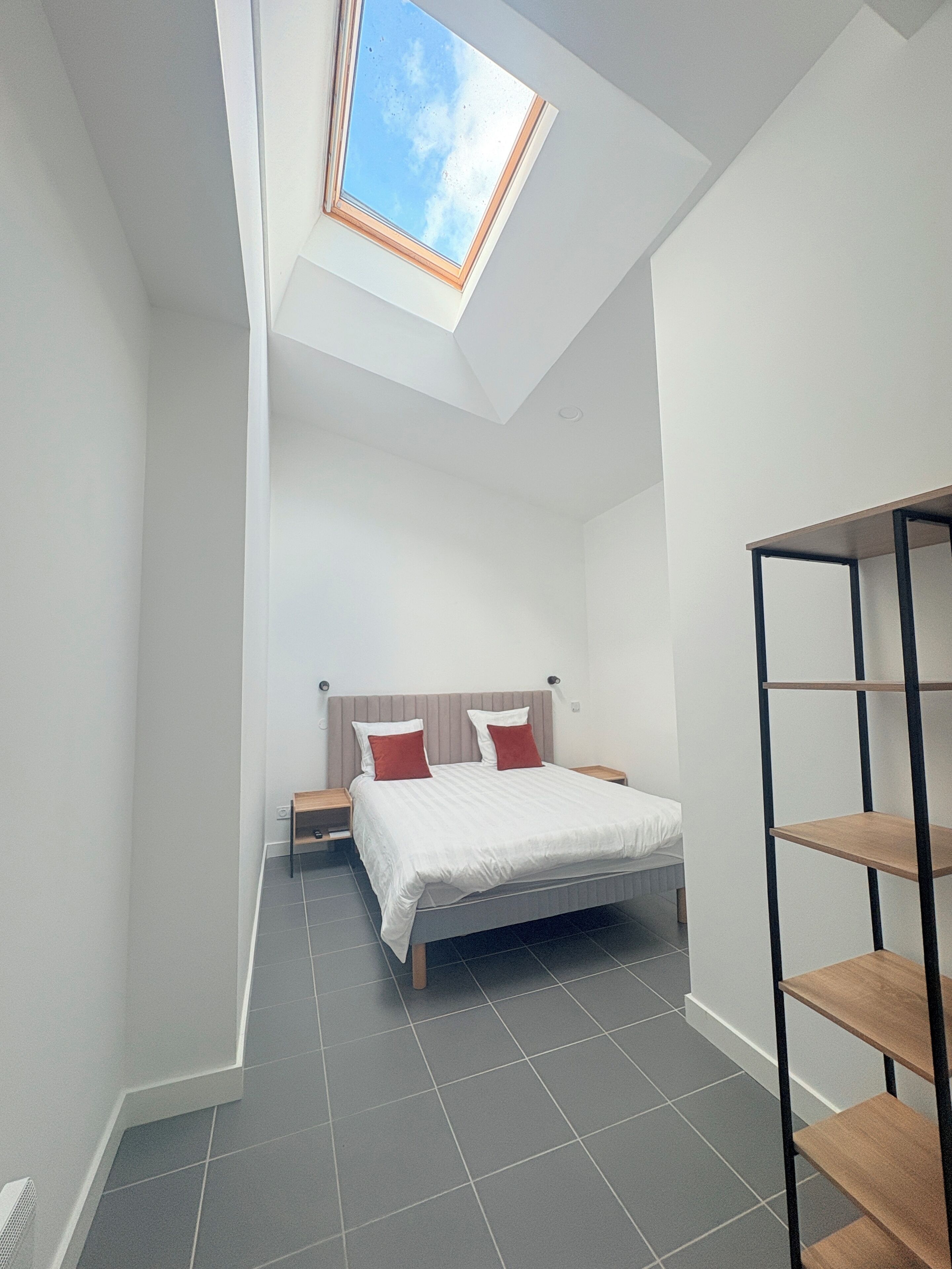 Une chambre avec un puits de lumière, un lit avec des oreillers rouges, une table de chevet en bois et une étagère en métal noir.