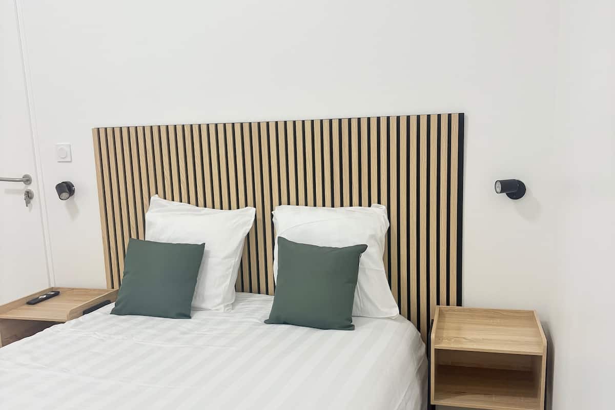 Un lit bien fait, avec une literie blanche et deux oreillers verts, une tête de lit en bois à rayures verticales et une petite table de chevet en bois avec une lampe.