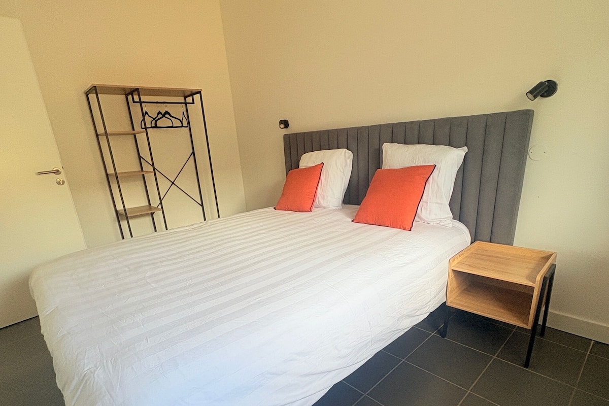Un lit bien fait, avec une literie blanche et deux oreillers orange, une table de chevet en bois et une étagère métallique adossée au mur.