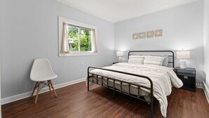 Un dormitorio con cama, mesitas de noche, una silla, una ventana y una lámpara en el techo.