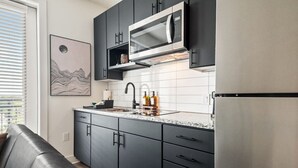 Eine moderne Küche mit dunklen Schränken, einem Edelstahlkühlschrank, einer Mikrowelle und einem Spülbecken mit Armatur. Auf der Arbeitsplatte liegen verschiedene Gegenstände, darüber hängt ein gerahmtes Kunstwerk.
