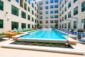 Une piscine dotée de chaises longues et d’un jacuzzi, le tout entouré d’un immeuble à plusieurs étages avec de grandes fenêtres.