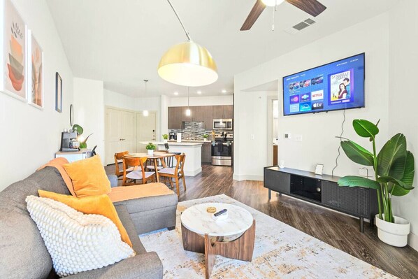 A modern living room with a gray sofa, orange cushions, a coffee table, and a TV displaying streaming services.