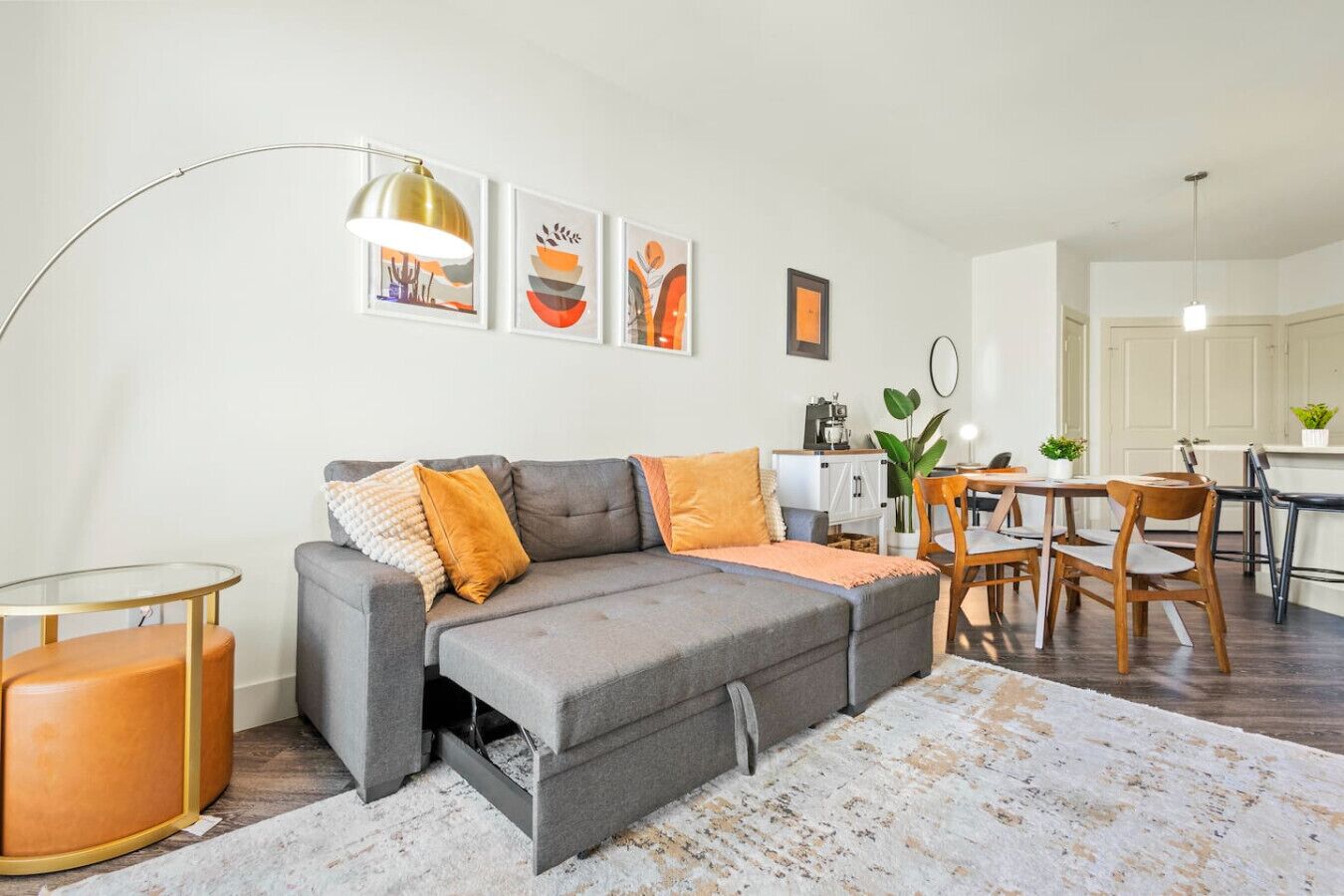 Un salon moderne avec un canapé d’angle gris, une table basse en verre et un coin repas avec des chaises en bois.