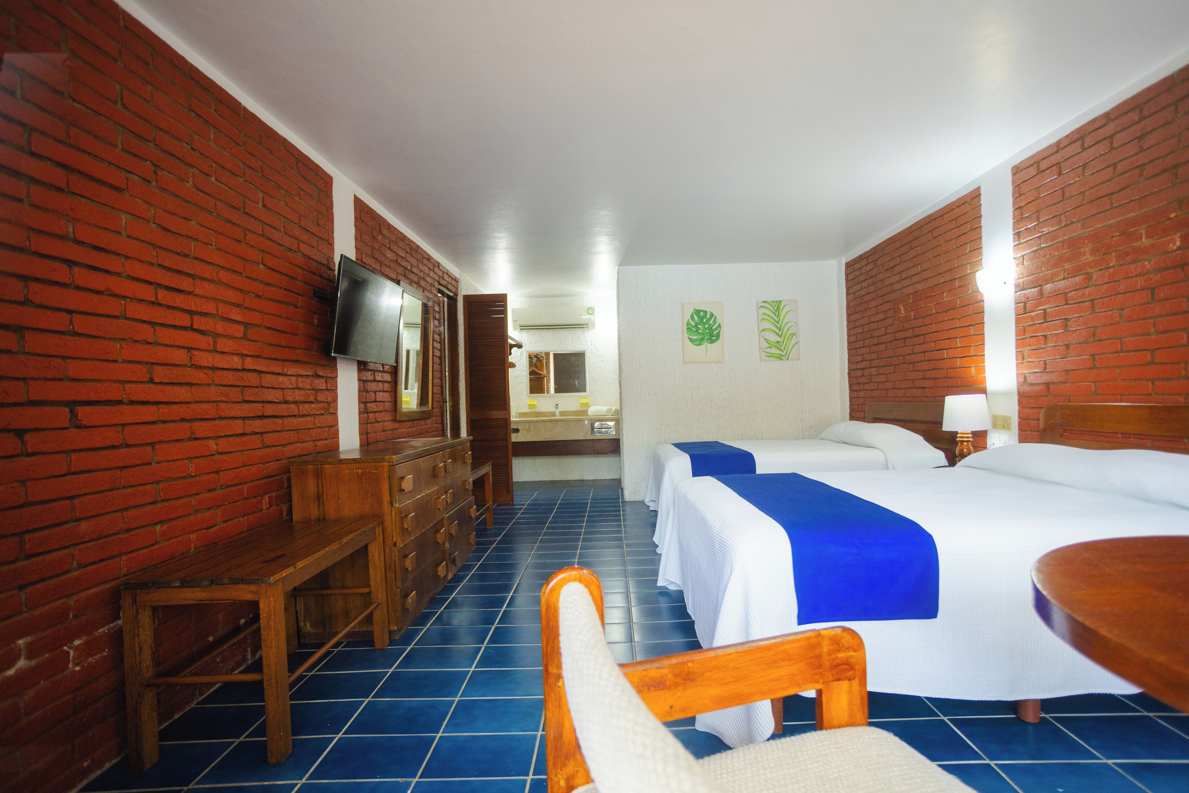 Une chambre d’hôtel avec un mur en briques, un téléviseur fixé au mur, un banc en bois, un bureau, une chaise, un lit avec une couvre-lit bleue et une table de chevet avec une lampe.