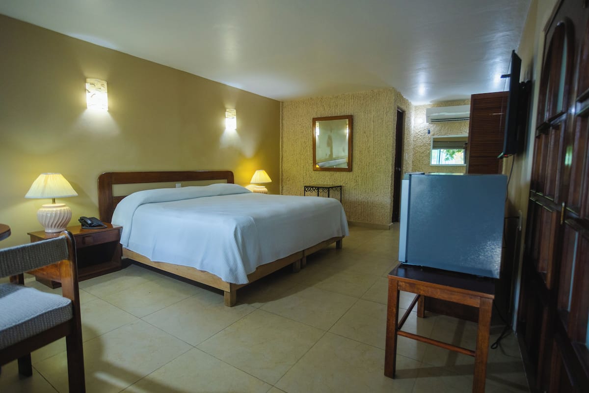 A hotel room with a bed, bedside lamps, a refrigerator, a chair, and a mirror on the wall.