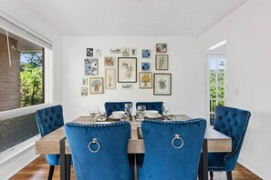 A dining room with a wooden table and blue upholstered chairs, framed artwork on the wall, and a view of greenery outside. - Orange Pit - 3BD 2BA Luxury Retreat - West Cola (West Columbia)