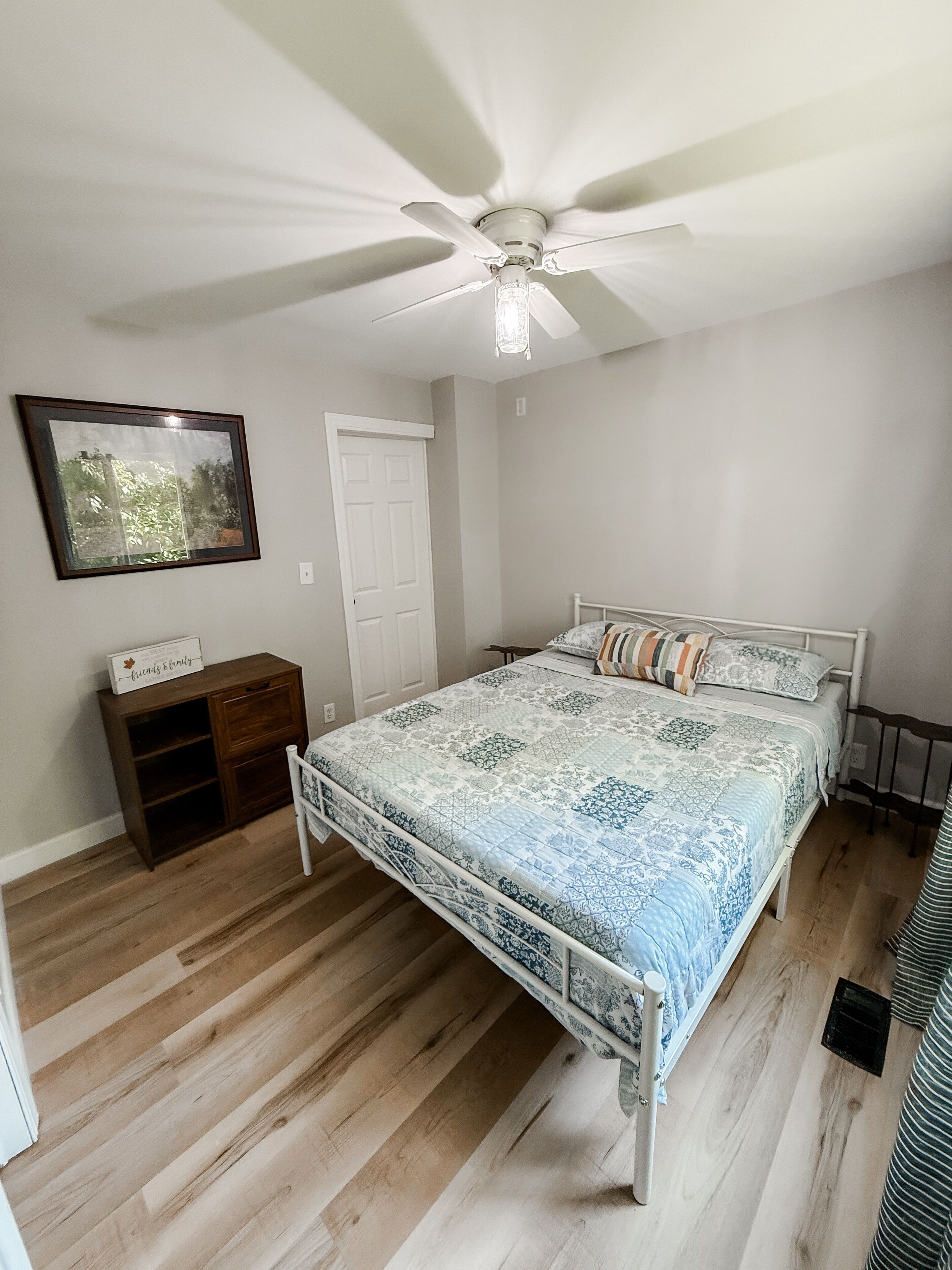 A bedroom with a bed, a ceiling fan, a wooden dresser, a framed picture, and a door.