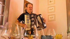 Un homme joue de l’accordéon dans une pièce dont les murs sont ornés d’œuvres d’art encadrées et où se trouvent des verres à vin sur la table.
