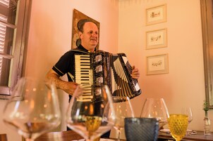 A man playing an accordion in a room with framed artwork on the wall and wine glasses on the table.