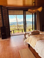A bedroom with a large bed, wooden flooring, a fireplace, and a view of a mountainous landscape.