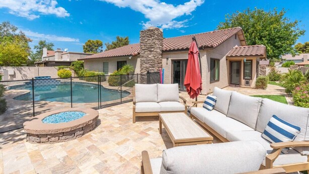 A poolside area with a stone fire feature, a seating area with a red umbrella, and a stone wall. - Sun, Fun & a Tempe Tan Home (Tempe)