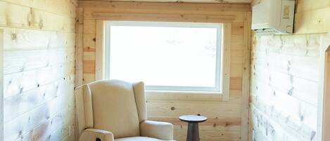 Ein kleiner, gemütlicher Raum mit Holzboden und -wänden, einem Fenster, einem beigen Sessel und einem kleinen runden Tisch.
