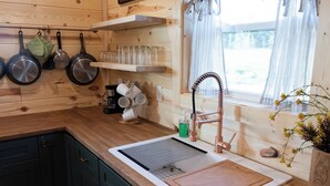 A kitchen with wooden cabinets, a sink with a cutting board, hanging pots, and a window with curtains.