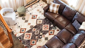 A cozy cabin interior with a brown leather sofa, a white chair, a TV mounted on the wall, and a patterned rug.
