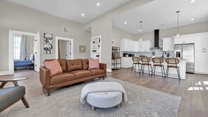 A spacious living room with a brown sofa, ottoman, and patterned rug. Adjacent is a kitchen with white cabinets, a stainless steel refrigerator, and bar stools. - Avondale Charmer-Comfort & Style (Birmingham)
