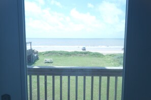 A view from a window overlooking a beach with a grassy area, a picnic table, and a vehicle parked on the sand.