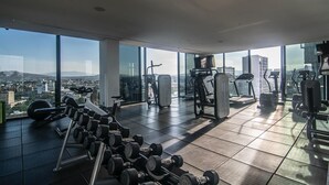 Un gimnasio en un edificio alto con diversos equipos de ejercicio y amplias ventanas con vistas a la ciudad.