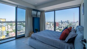 Un dormitorio moderno con una cama grande, un televisor y vistas a un paisaje urbano a través de ventanas de piso a techo.