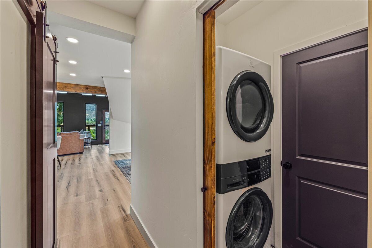 A laundry room with two stacked front-loading washers and dryers, a wooden door, and a view into a living area with a couch and a dining table.