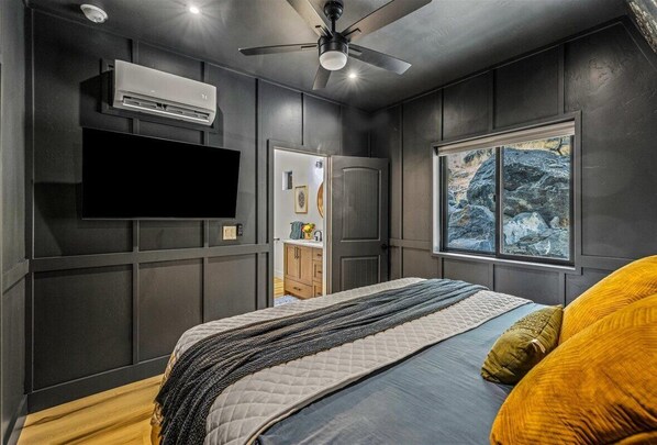 Un dormitorio con cama, ventilador de techo, unidad de aire acondicionado, ventana con vista a rocas y puerta que da a un baño.