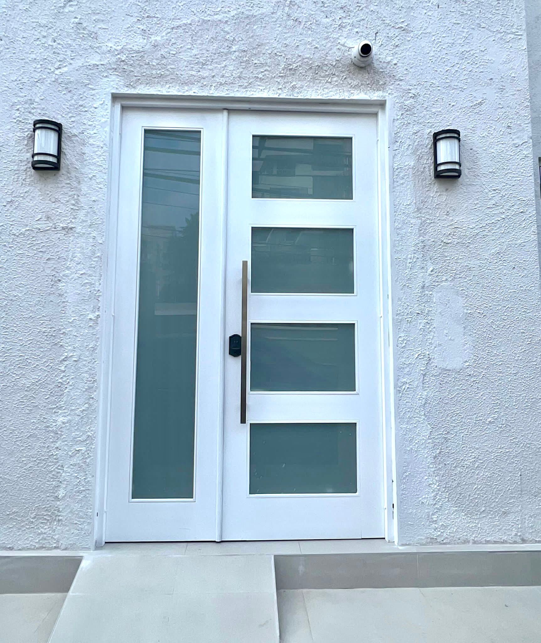 Una puerta doble blanca con paneles de vidrio esmerilado y un pomo negro, flanqueada por dos apliques de pared.