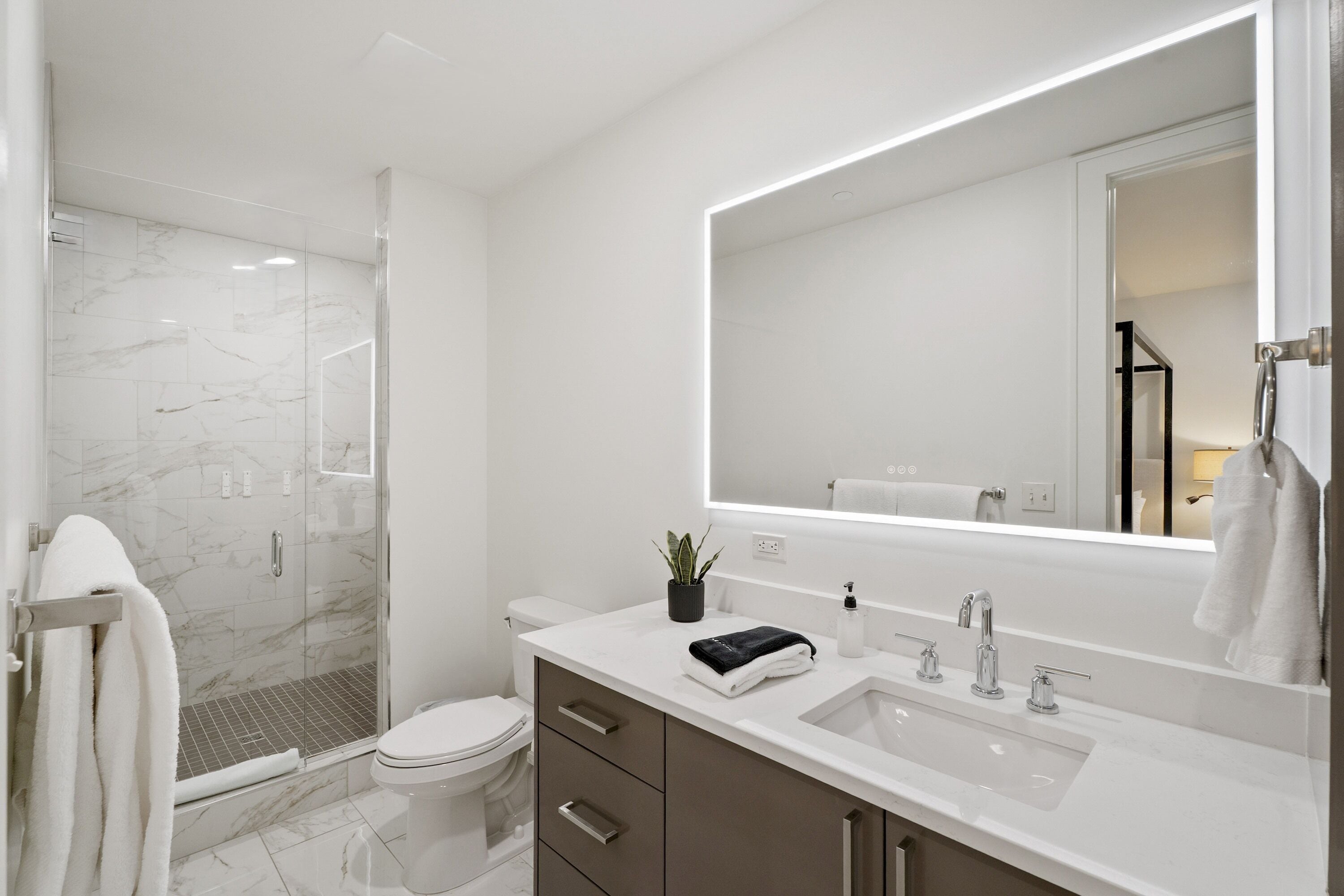 Un baño moderno con cabina de ducha de vidrio, inodoro blanco, espejo grande y lavamanos blanco con toalla negra.