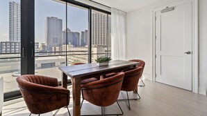 A modern dining area with a wooden table and four brown chairs, large windows offering a city view, and a white door. - The Theodore 3BD/3B - All King Bedrooms (New Orleans)
