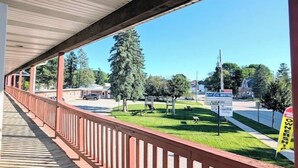 A covered outdoor area with a wooden railing, a clear view of a motel and a church in the background, and a grassy area with trees. - Cool City Motel  (Two Rivers)
