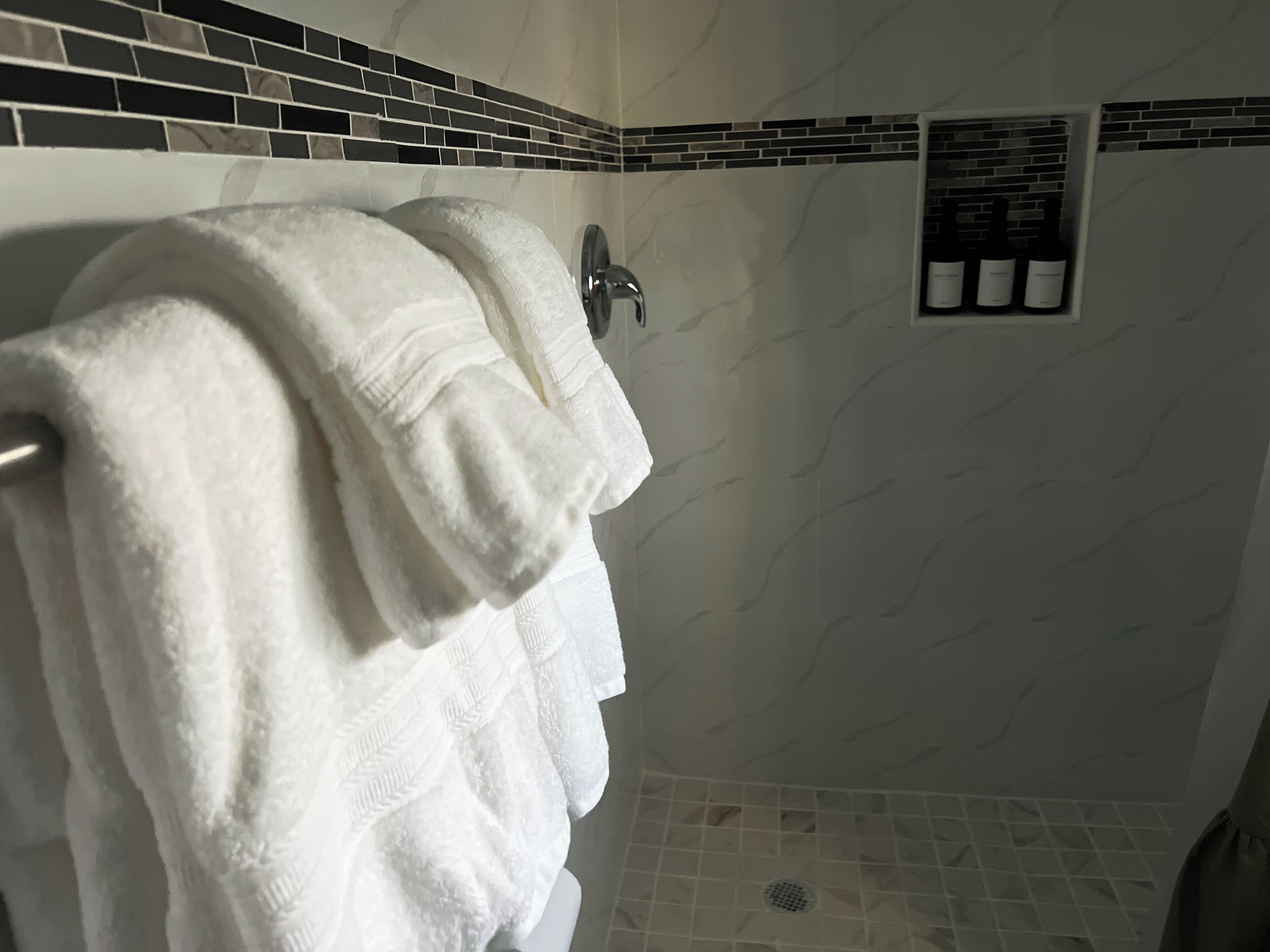 A bathroom with a towel rack holding multiple white towels, a showerhead, and a tiled wall with a black and white pattern.