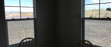 A corner room with a table and two chairs, large windows with a view of a gravel area and distant landscape.