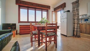 A modern kitchen with wooden cabinets, a dining table with chairs, a refrigerator, and a large window with curtains. - Bongiovi Houses - App. Azure - 2 min from the beach, with garden and sea view (Sicilia)