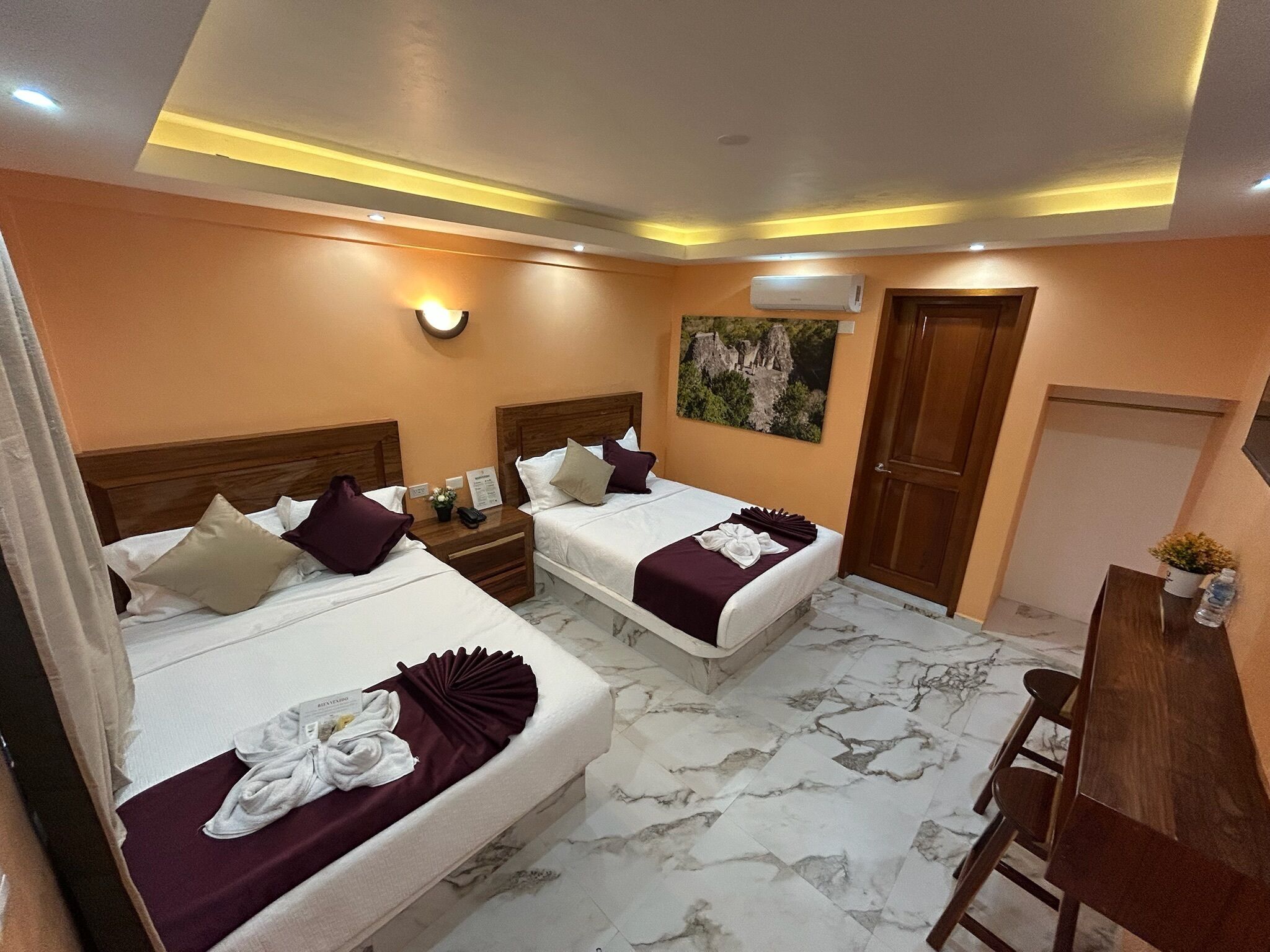 Habitación de hotel con dos camas, un escritorio, una silla, una mesita de noche, aire acondicionado instalado en la pared y un cuadro.