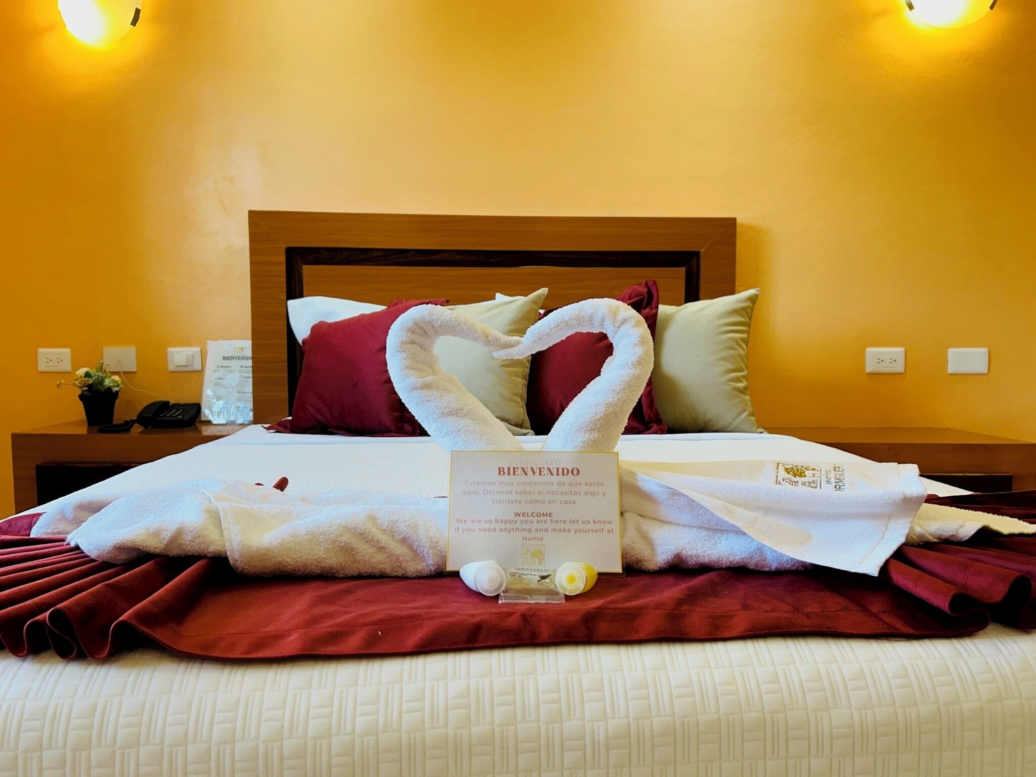 A hotel room with a neatly made bed, a welcome sign, and a wall-mounted light fixture.