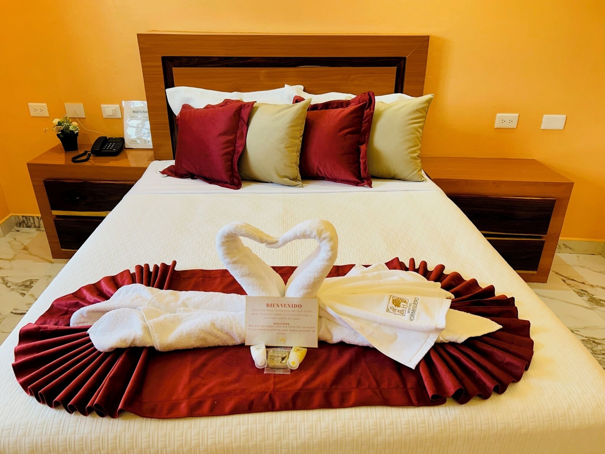A neatly made bed with a heart-shaped towel arrangement, a welcome card, and a phone on the bedside table.