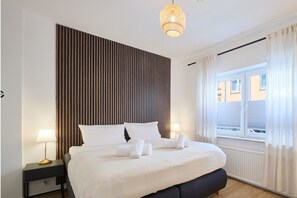 A hotel room with a large bed, two bedside tables, a hanging light fixture, a window with curtains, and a view of buildings outside. - NUIT Apartment | Modern Flat in the city (Krefeld)