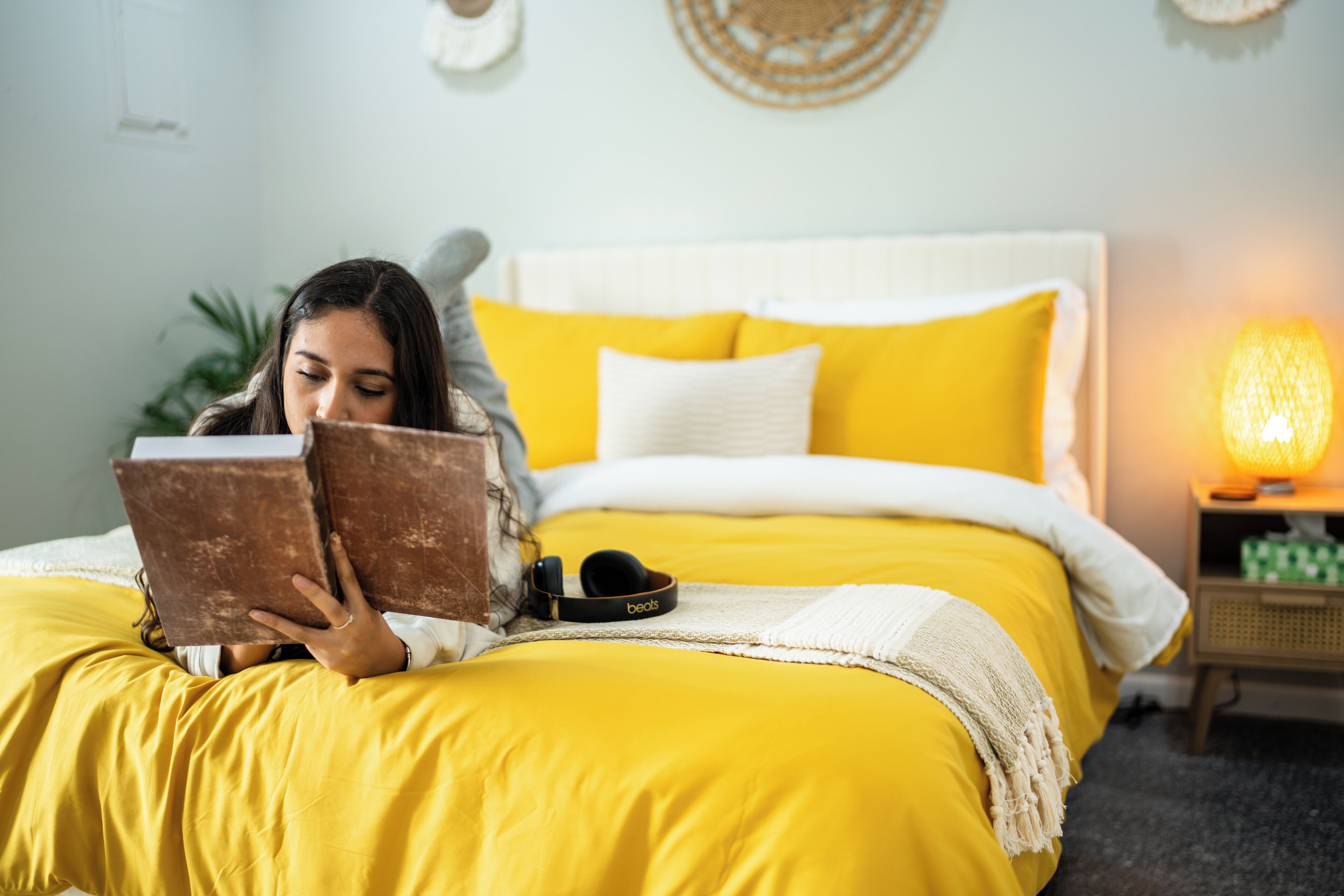 Iemand ligt in bed met geel beddengoed, een boek lezend.