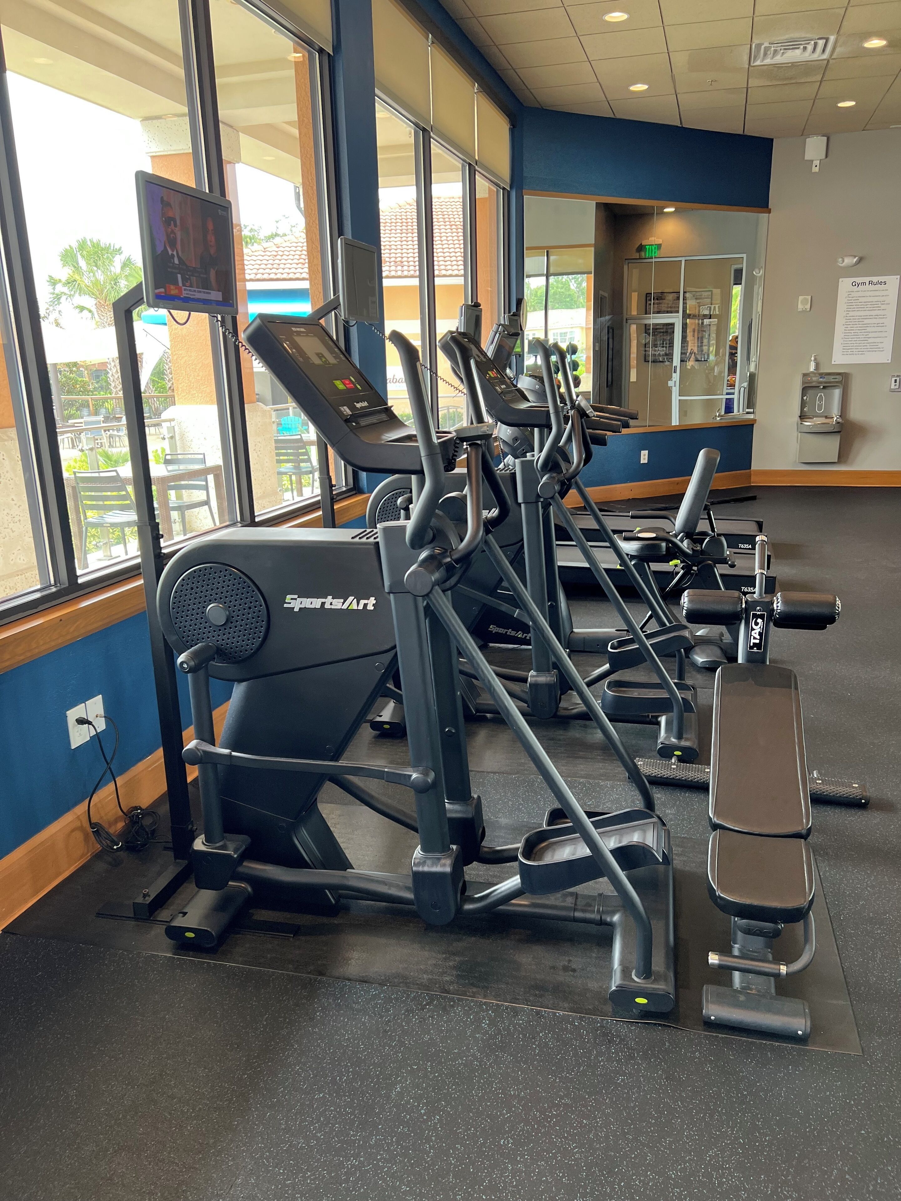 Une salle de sport équipée de nombreux appareils d’exercice, dotée de grandes fenêtres et d’un distributeur d’eau.