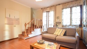 A living room with a wooden staircase, a sofa with a striped pillow, a coffee table with a bowl and a magazine, and large windows with floral curtains. - Lovely home in Seravezza with WiFi (Seravezza)