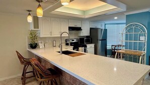 Una cocina con una isla grande, armarios blancos, un refrigerador de acero inoxidable y una zona de comedor con una mesa y una silla de metal.