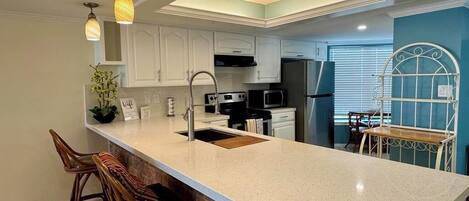 A kitchen with a large island, white cabinets, a stainless steel refrigerator, and a dining area with a metal chair and table.